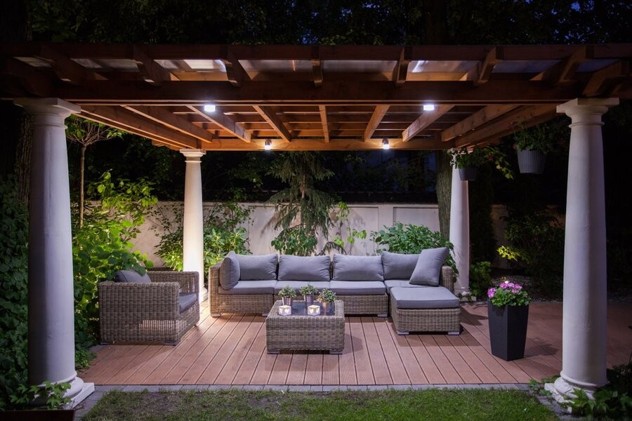 An outdoor patio setup under a pergola with seating and outdoor lighting fixtures.