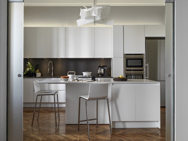 Modern kitchen with a minimalist design featuring white cabinetry, a central island with bar stools, built-in stainless steel appliances, and warm wooden parquet flooring.