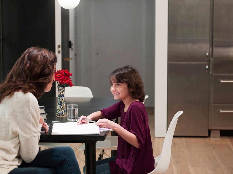 A mother and daughter sit at a table in a modern kitchen, smiling and conversing as the daughter does homework.