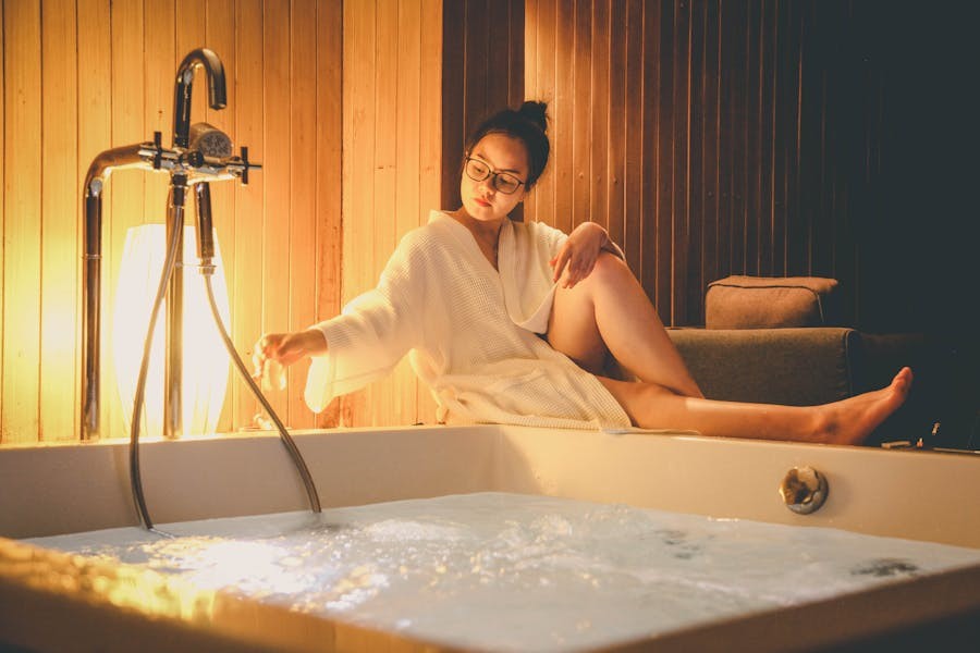 Woman wearing a white bathrobe sitting beside a white bathtub filled with water.