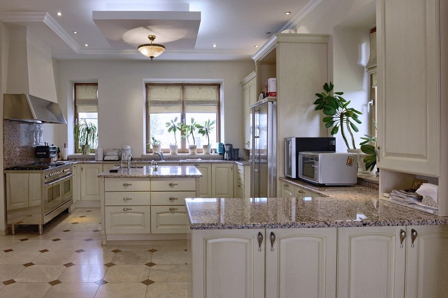 A kitchen space featuring ceiling lighting fixtures and motorized shades on windows in the background.